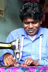 Bangladeshi man wearing a blue shirt looking down and using sewing machine with small motor attached.