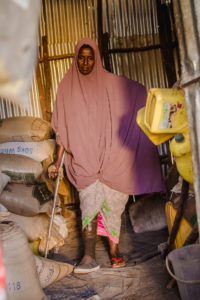 Woman wearing pink hijab with a prosthetic leg and a crutch walks through her store with cereals on her left and cooking oil cans on her right. The store is made of corrugated metal and tree branches.