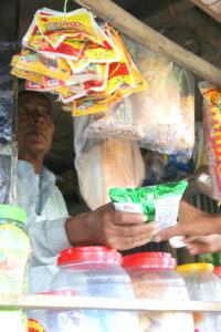 6: A blind shopkeeper gives a bag of chips to a customer at front counter of shop, there are storage conatiners of various colors, sizes and texture in front of shop.