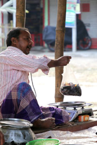 Bangladeshi man sitting on a platform in a market with several male customers standing around him, holding a bag of fish over an electronic scale.