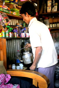 A shop keeper is standing with a kettle in hand and pouring tea. Canned items are on shelves around him.