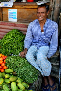 A man with a blue striped collared shirt and blue and white pants sits along the side of a bed of vegetables while smiling.