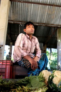 Man sits on a stool near vegetables he is selling.