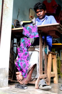 A tailor put his feet one on top of the other on a foot pedal to activate the motor of his sewing machine to sew a garment.