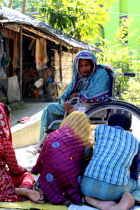 Woman in a wheelchair teaches 6 children seated on the ground. Her home, which has an accessible ramp, is in the background.