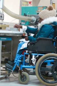 A woman is seated in a power wheelchair in front of a high table. She reaches for a plastic part to put into a small bag for an assembly line.