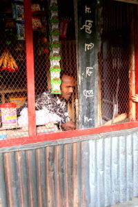 A man sits in a shop behind a wire mesh covering the front. To the right of him, a small girl customer leans over to purchase an item.