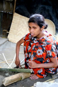 A woman sits in front of a half bamboo log, a sickle on her right and strips of bamboo on her left.