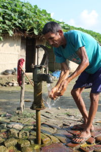 A man is stooped close to a water pump, he is washing his hands while water flows. His walking stick is rested on a post behind him and his house can be seen in the background.