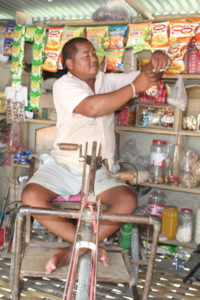 Man who uses a tricylce is sitting in his shop, surrounded by items to sell, he tears a snack off a stringed line.