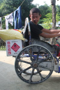 Man sits on tricycle in his home, with boxes and canvas bags secured on the back and side of his tricycle.