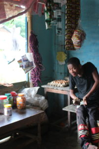 Man sits inside his shop, holding cash he is exchanging with a female customer standing at front window. He has bottles of candies and eggs around him.