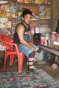 Man sits inside shop, wearing bilateral prosthetics, surrounded by his good to sell. A colorful carpet is on the ground.