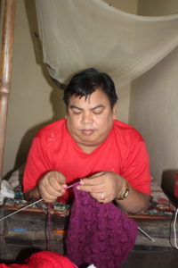 Man knits a purple garment, body propped up at the end of his bed.