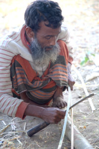 Man splits a piece of bamboo in a squat position using a sickle.