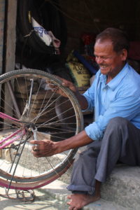 Man sits on the steps of his shop, using a wrench to repair the back tire of a bike that is upside down.