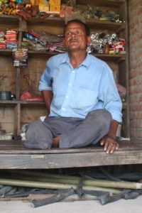 Shopkeeper sits on a table with tire tubs under the table and shelves behind him with various items.