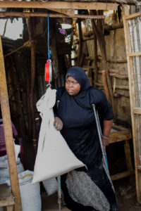 Woman stands using crutches and is weighing a canvas bag filled with food in her right hand, and holding a crutch with her left hand.