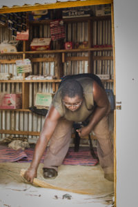 Man is bent forward sweeping the floor of his shop, he is wearing leather shoes.
