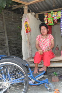 Woman sits inside her small shop, with a blue wheelchair on the left, and her items for sale on her right on the floor of the shop. Additional items hang on an overhead string.