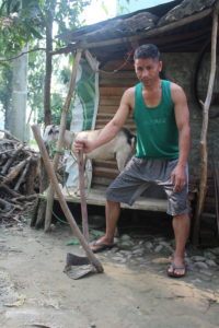 Man stands with knees bent and holding hoe/axe tool used for farming. A goat stand behind him on a wooden shack.