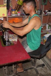 Man sits in his shop with items and electronics behind him. He is sitting a table and threading a sewing machine. He is rotated slightly to the right and sitting on a swivel chair.
