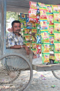 A profile view of a man sitting in his tricycle with many differently colored packets hanging from the roof of his tricycle.