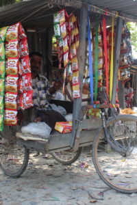 A front view of the mobile-tricycle shop the man runs, with items contained on the floor of the tricycle and hanging off the roof built above the tricycle.