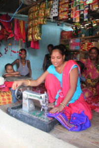 Female tailor sits in front of her sewing machine on raised wooden floor of shop, she uses her right hand to turn the wheel of the machine. She is surrounded by sales items.