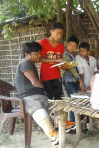 A man with two prosthetic legs is seated on a chair. He is watching 3 boys read their school work, while six other children sit in front of him on a short bamboo table reading their homework.